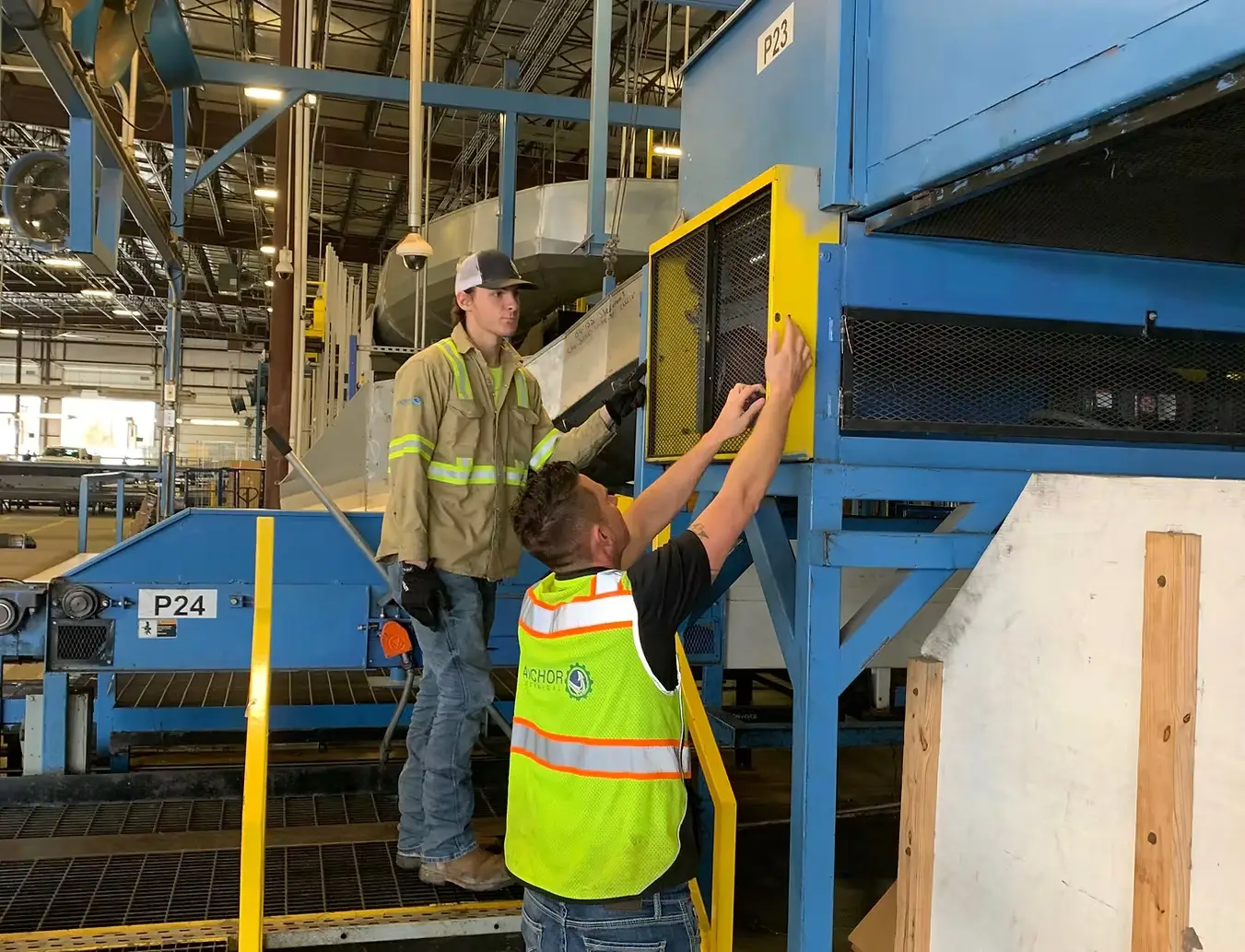 Industrial Workers Performing Machine Maintenance in Factory Two industrial workers inspecting and maintaining machinery in a factory setting, wearing safety gear and reflective vests